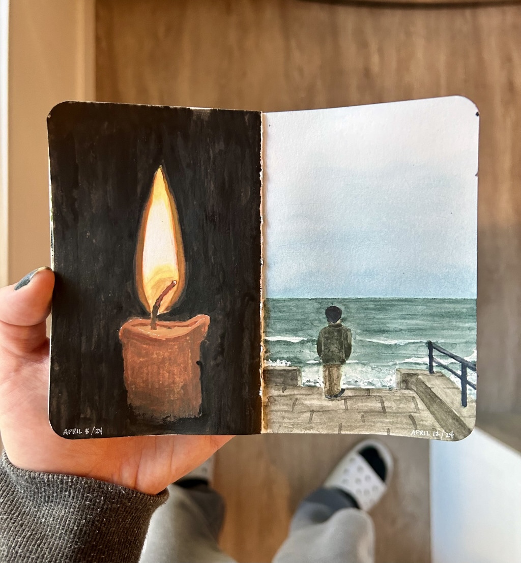 A small sketchbook being held open to show two paintings, on the left a lit candle against a black background and on the right, a figure looking out over the sea on an overcast day