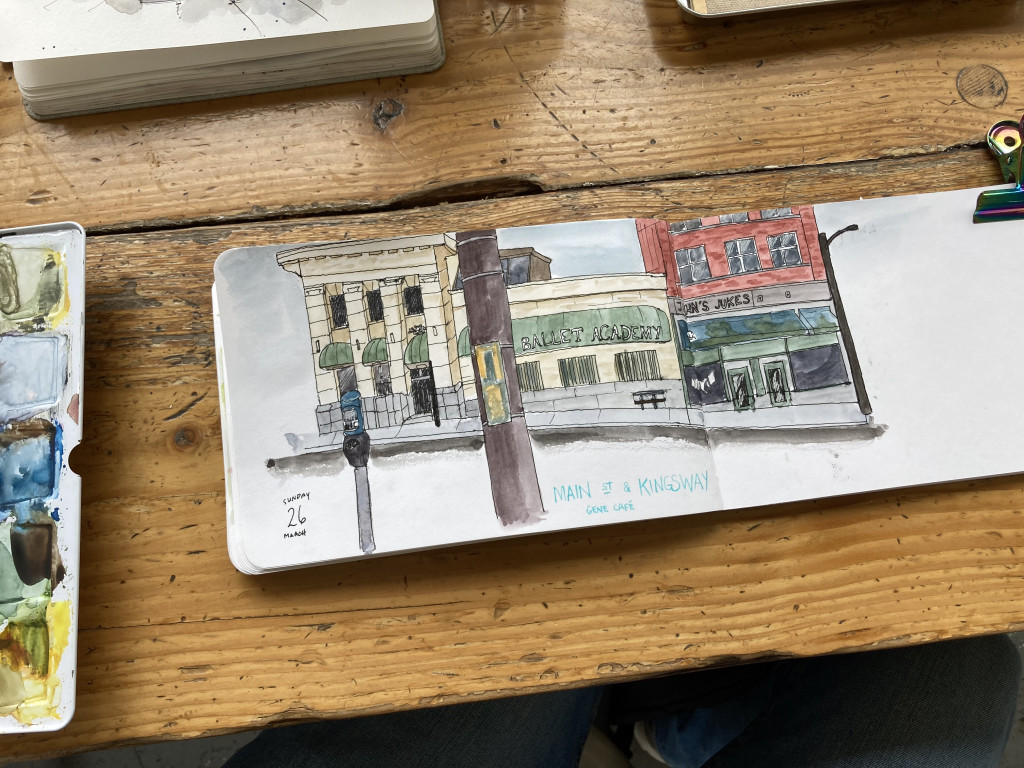 An ink and watercolour sketch in a sketchbook of three buildings as seen from across the street, labelled Main Street and Kingsway, Gene Cafe. Next to the sketchbook is the edge of a paint palette, and the edge of another sketchbook.